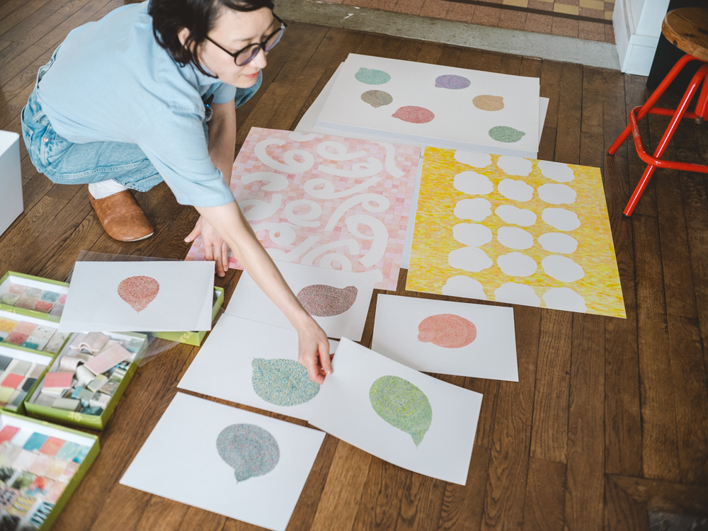 A photo of Yoko Homareda arranging her ceramic-inspired graphic prints on a wooden floor, showcasing colorful textured patterns and speech-bubble shapes alongside her art supplies.